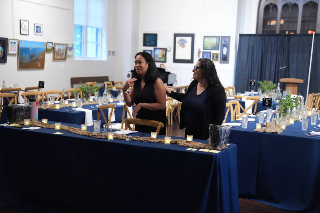 Maricar stands behind a table setting communicating in ASL with an interpreter by her side to a group off camera.