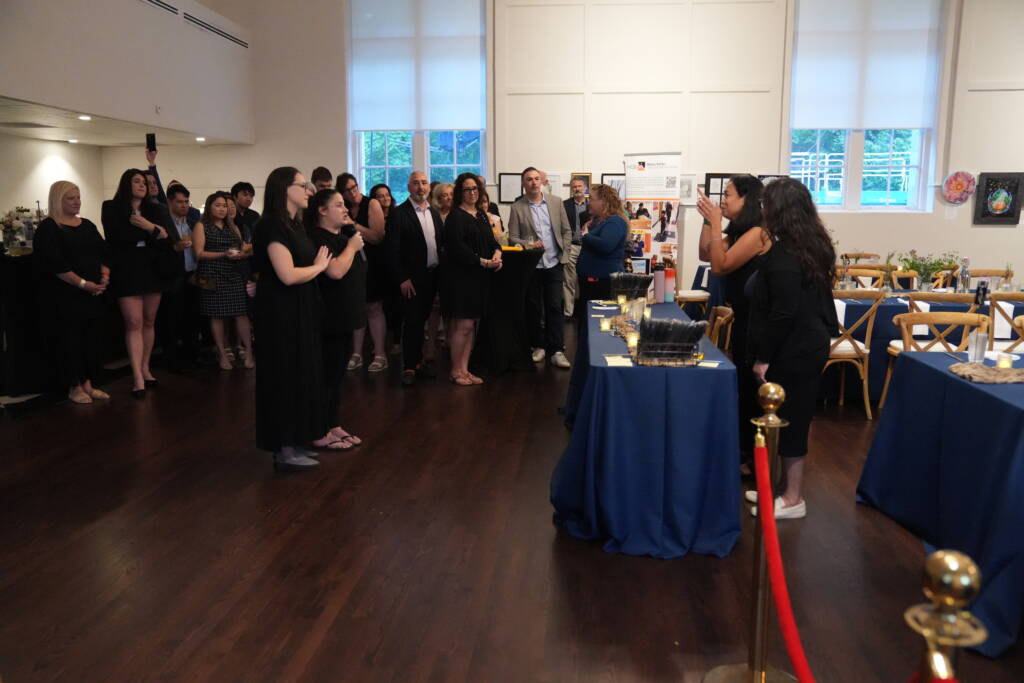 Maricar stands behind a table setting presenting in ASL with an interpreter by her side to a large group of guests standing in front of her   