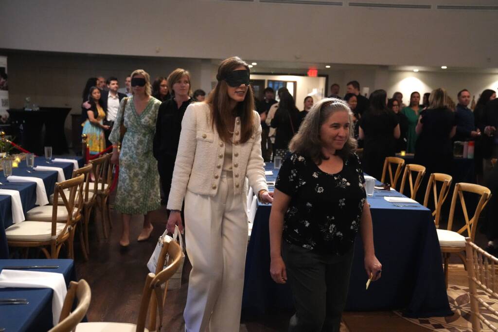 A female guest is guided under blindfold to her table by an HKS staff member, while others follow behind them.