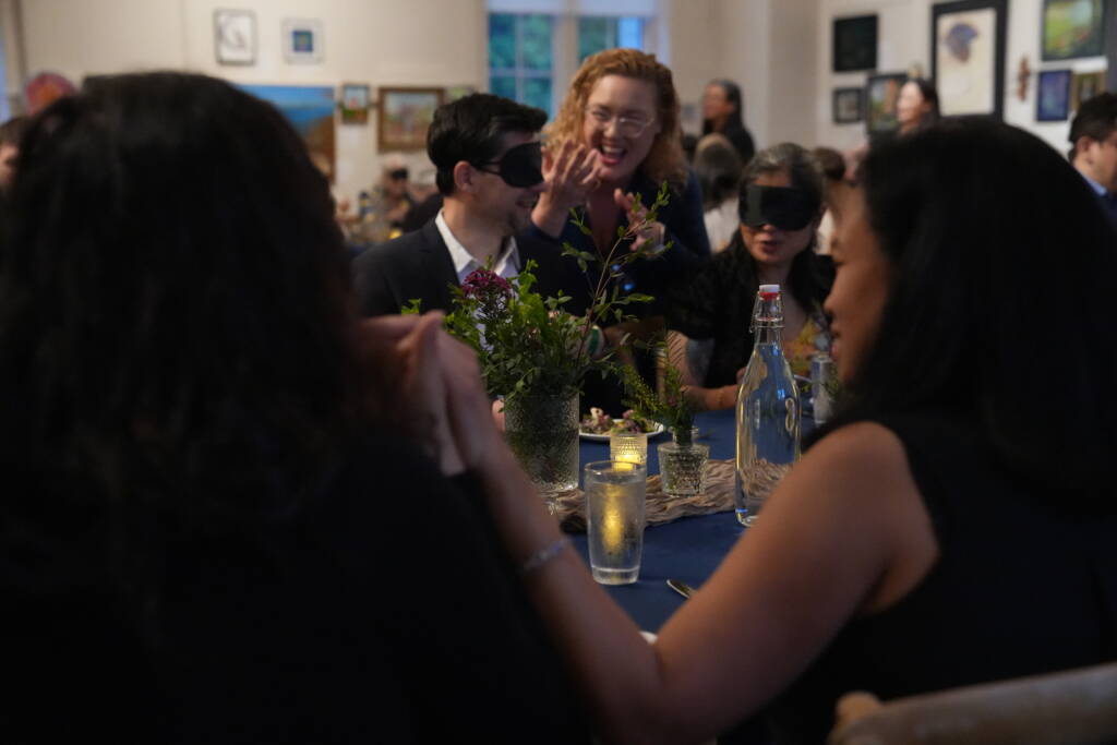 An interpreter is signing in ASL behind a guest wearing a blindfold.  In the foreground is Maricar and an interpreter communicating in tactile ASL.   