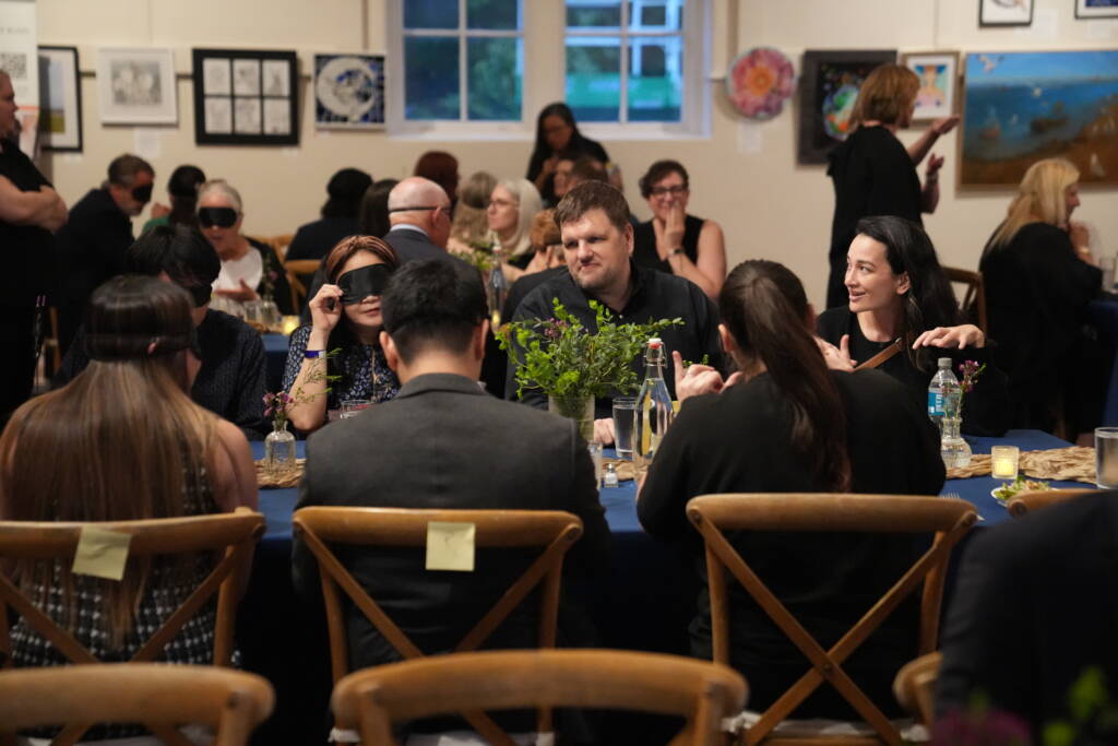 Ryan is sitting at a table alongside an interpreter and they are engaging in conversation with guest wearing blindfolds who are sitting with them. 
