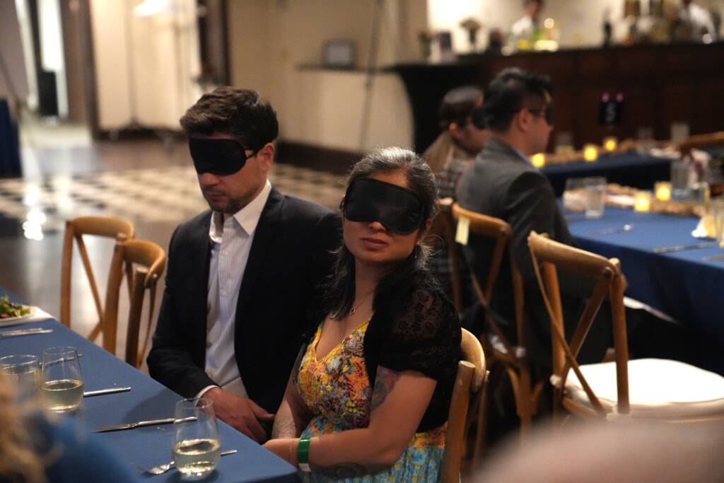 A man and a woman sit together both wearing blindfolds. 