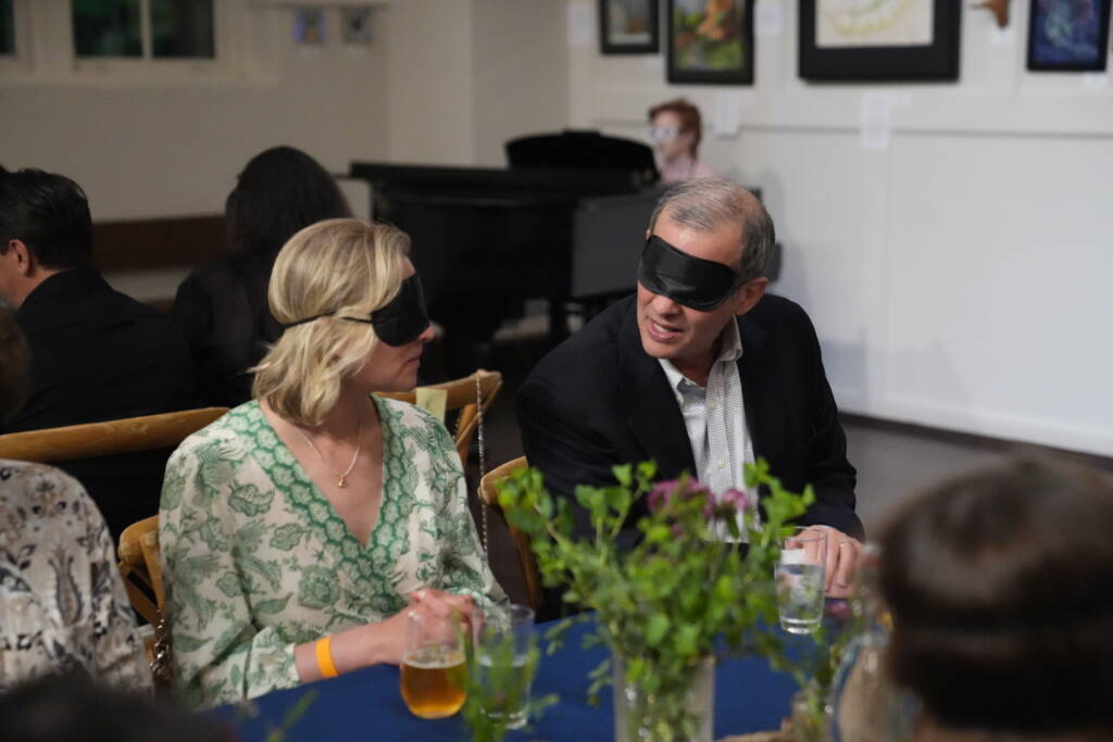 A man and woman both wearing blindfolds engaging in conversation while seated at a table. 