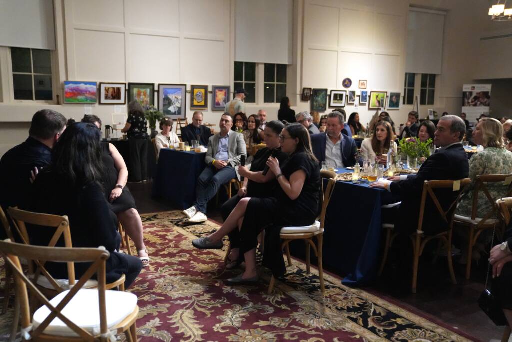 Guests who are seated at several tables look on as Ryan and Ashley have their discussion. Two interpreters holding microphones are sitting and facing Ryan and Ashley.  