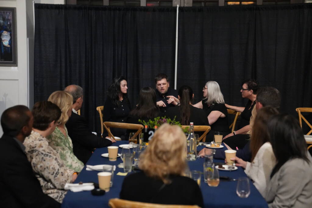 Ryan Odland communicating in ASL to a group with Ashley by his side. Her had is placed over his.  A table of guests are facing Ryan and Ashley.  