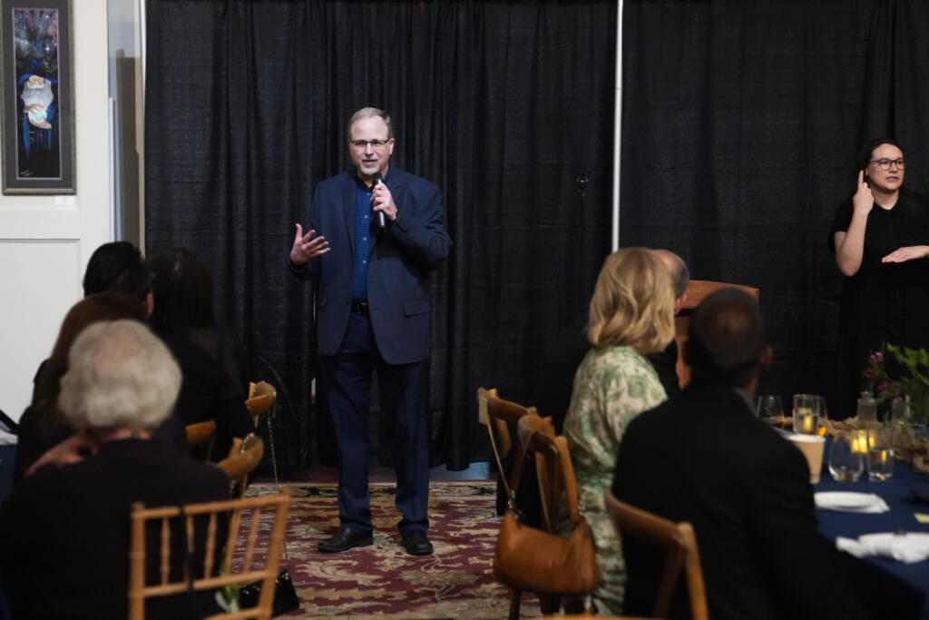 Jeff Kunkel is standing and holding a microphone while speaking in front of event attendees.  