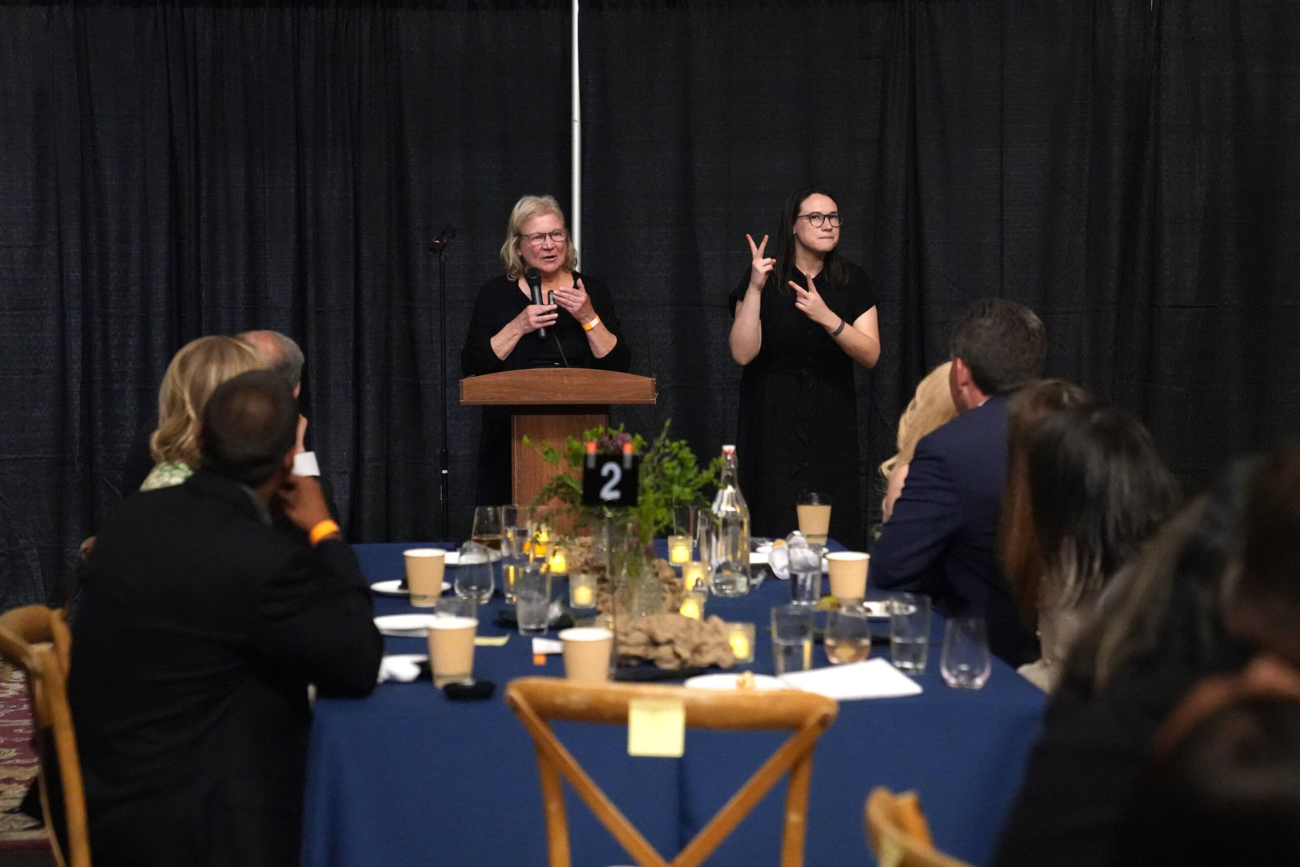 Sue Ruzenski is standing at a podium holding a microphone and speaking to guests with an interpreter standing to her left signing in ASL.  