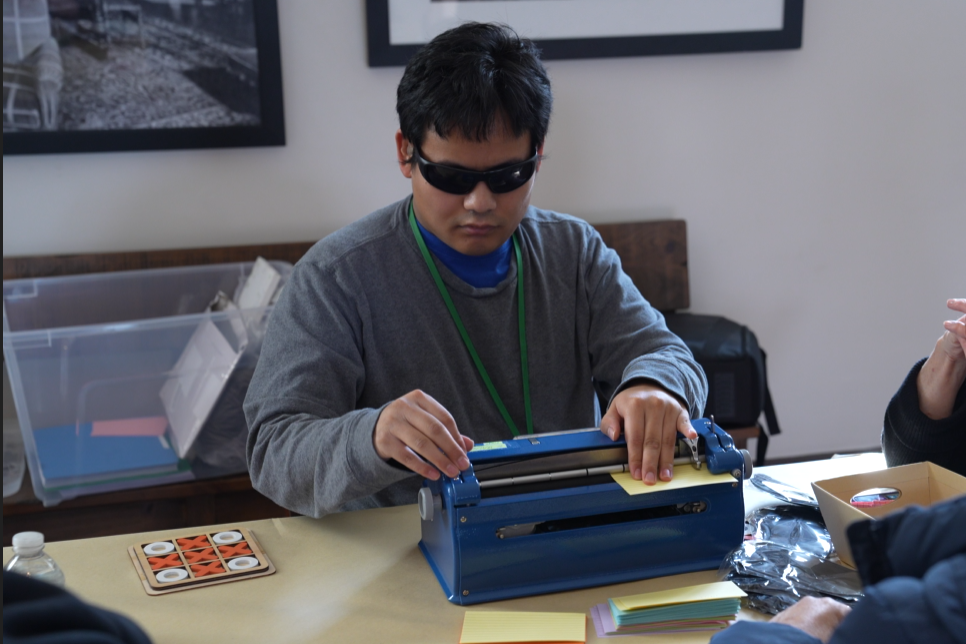 A young adult wearing sunglasses works on a Perkins brailler.