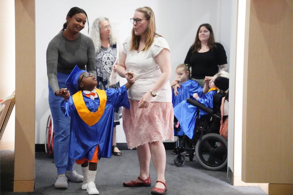 Preschoolers wearing blue and gold graduation gowns and caps stand with teachers.