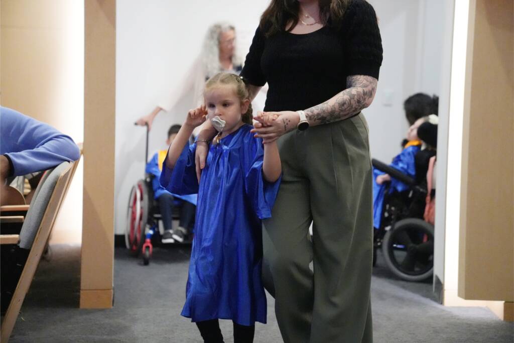 A Preschool girl wearing a blue graduation gown walks with a teacher.