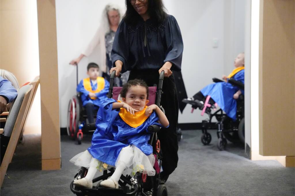 A preschool girl in a mobility stroller wearing a blue graduation gown enters the ceremony.