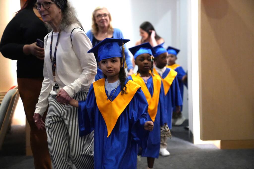 Preschoolers wearing blue caps and gowns walk alongside staff members as the enter the graduation ceremony.