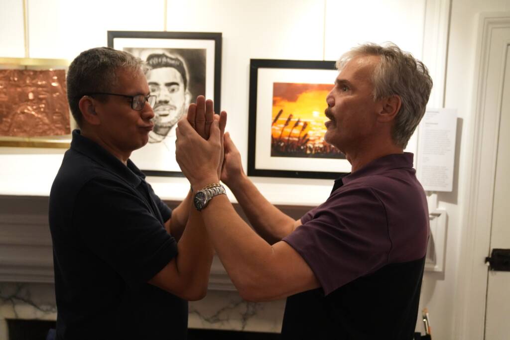 Two individuals communicating in tactile ASL while standing in front of artwork. 