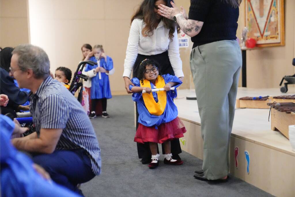 A preschool girl wearing a blue and gold graduation gown holding a certificate stands with a teacher and smiles.
