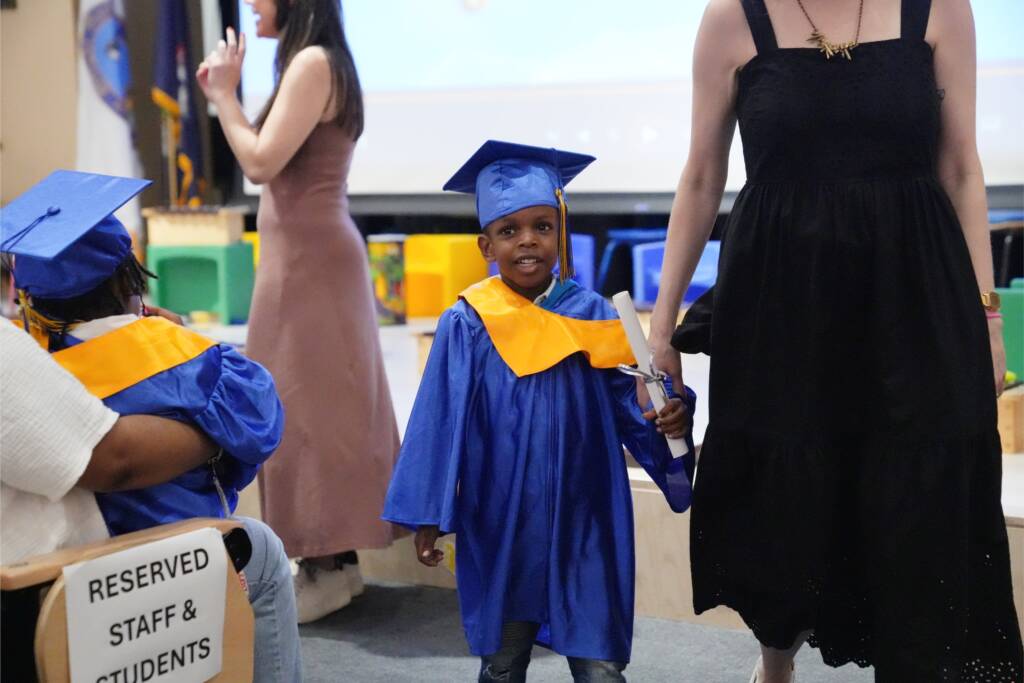A preschool boy girl wearing a blue graduation gown and cap and holding a certificate walks with a teacher.