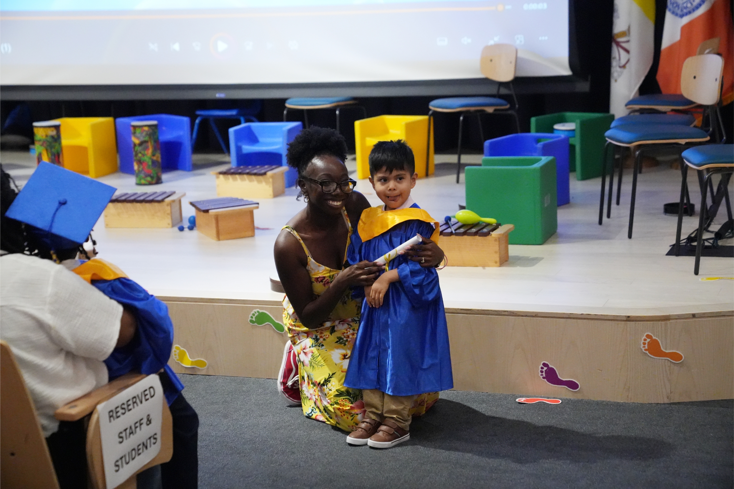 A preschool boy wearing a blue and gold graduation gown stands with an CLC staff member while receiving a certificate.