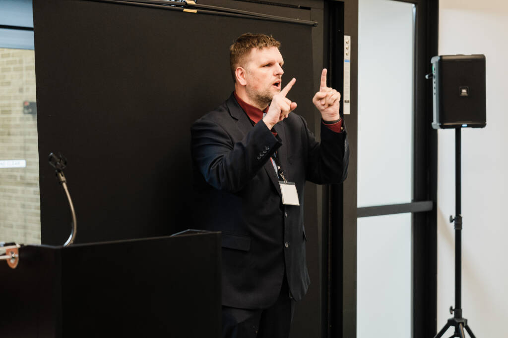 Ryan Odland signs in ASL while standing in front of a black backdrop positioned in the atrium.