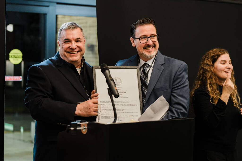 Mayor Peter Forman and John Filek stand side by side, both holding a framed proclamation between them. A sign language interpreter is visible on the right edge of the frame