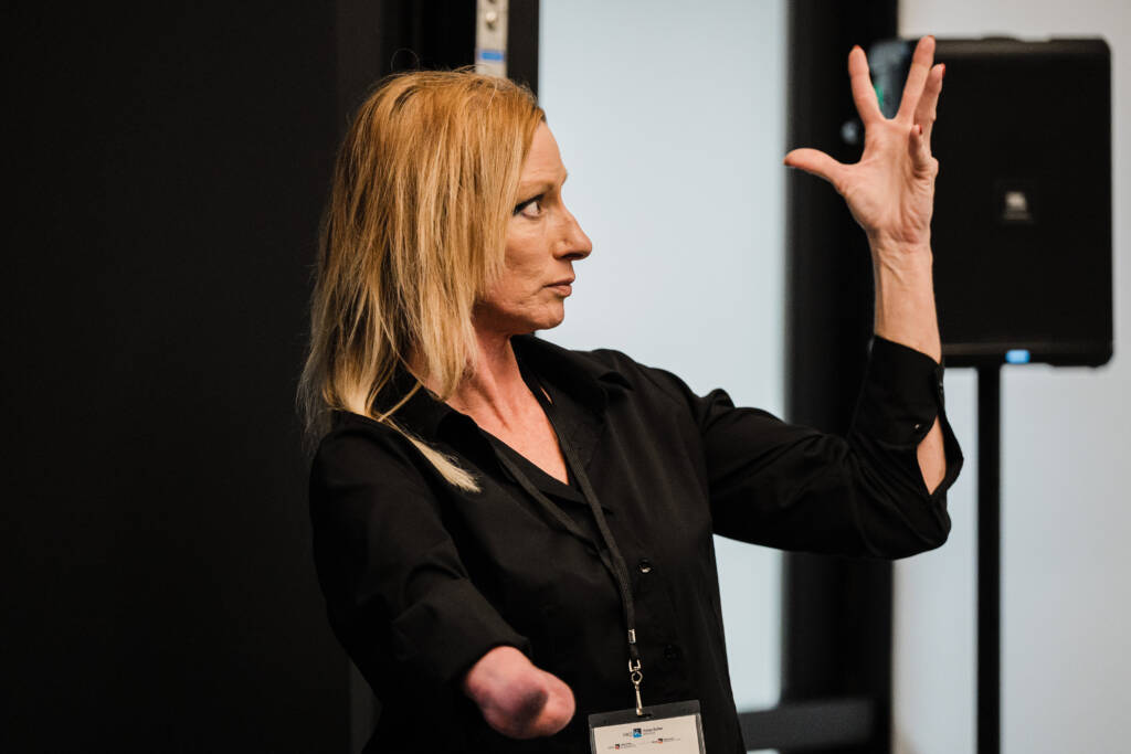 Elizabeth Schmaltz communicating in sign language, her left hand raised with fingers extended. She wears black professional attire and has shoulder-length blonde hair.