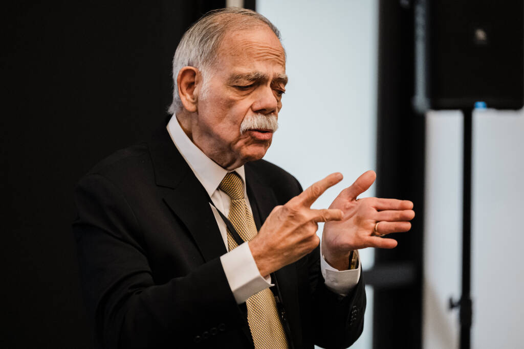 Dr. Francis Casale signs to event attendees, both hands positioned expressively in front of his chest. He wears a dark suit with a white shirt and gold patterned tie.