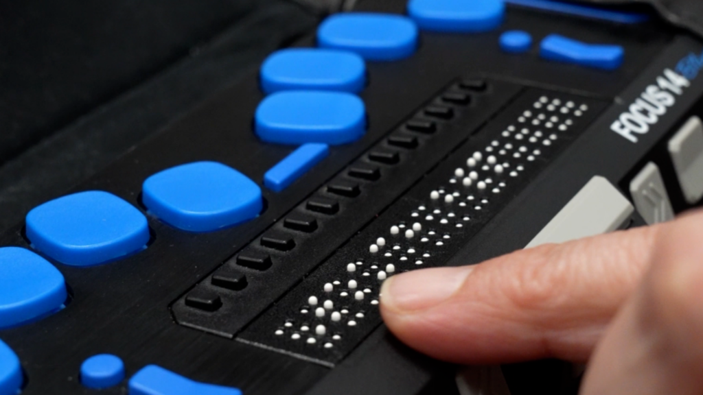Finger reading refreshable braille display with raised pins and blue navigation buttons.