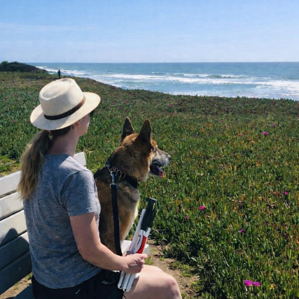 Woman in a sun hat sits on a bench with a German Shepherd, overlooking coastal flowers and the ocean