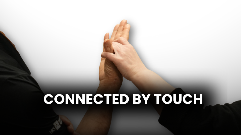 Two hands connect in tactile sign language at the center of the frame, one palm open and facing inward with the other hand lightly interlaced to receive the signs. Bold white text reads: CONNECTED BY TOUCH.