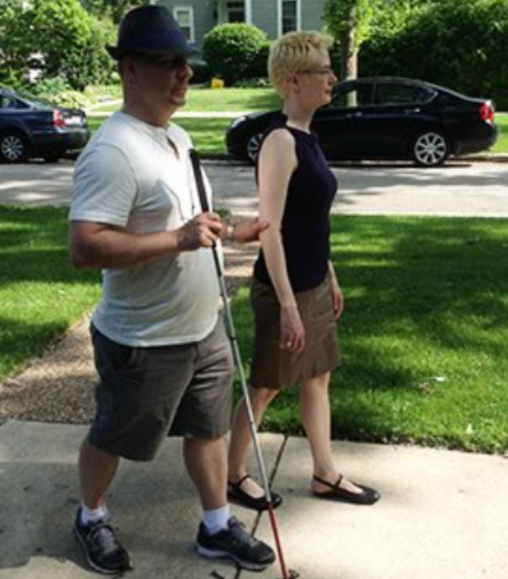 A man holding a white cane in one hand and the arm of a sighted guide, walking together on a sidewalk.