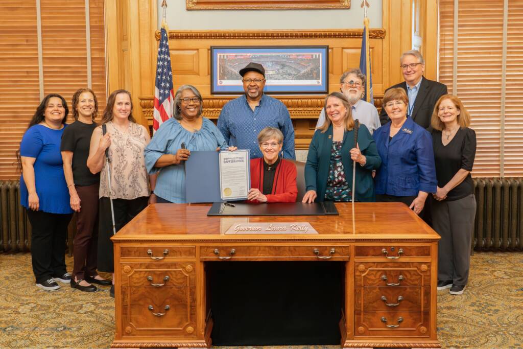 Kansas Governor Laura Kelly holds the signed 2026 DeafBlind Awareness Week proclamation, surrounded by DeafBlind Kansans, family members, and HKNC staff.
