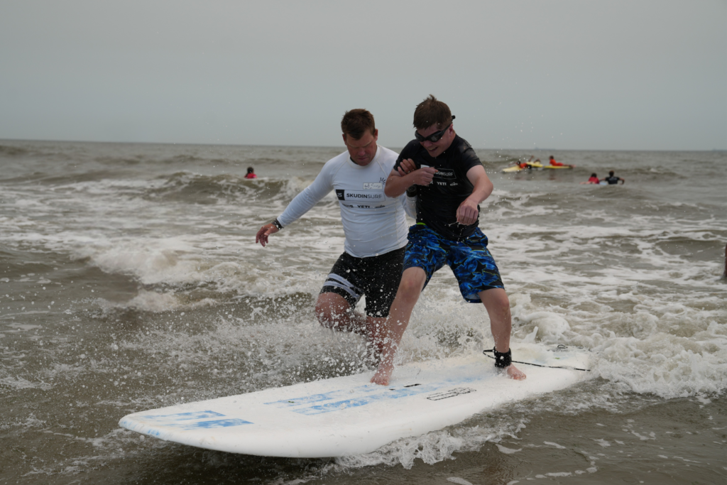 A Summer Youth Vocational Program participant stands on a surfboard riding whitewater toward shore, smiling, as a Skudin Surf instructor wades close behind with a supporting hand on his arm.