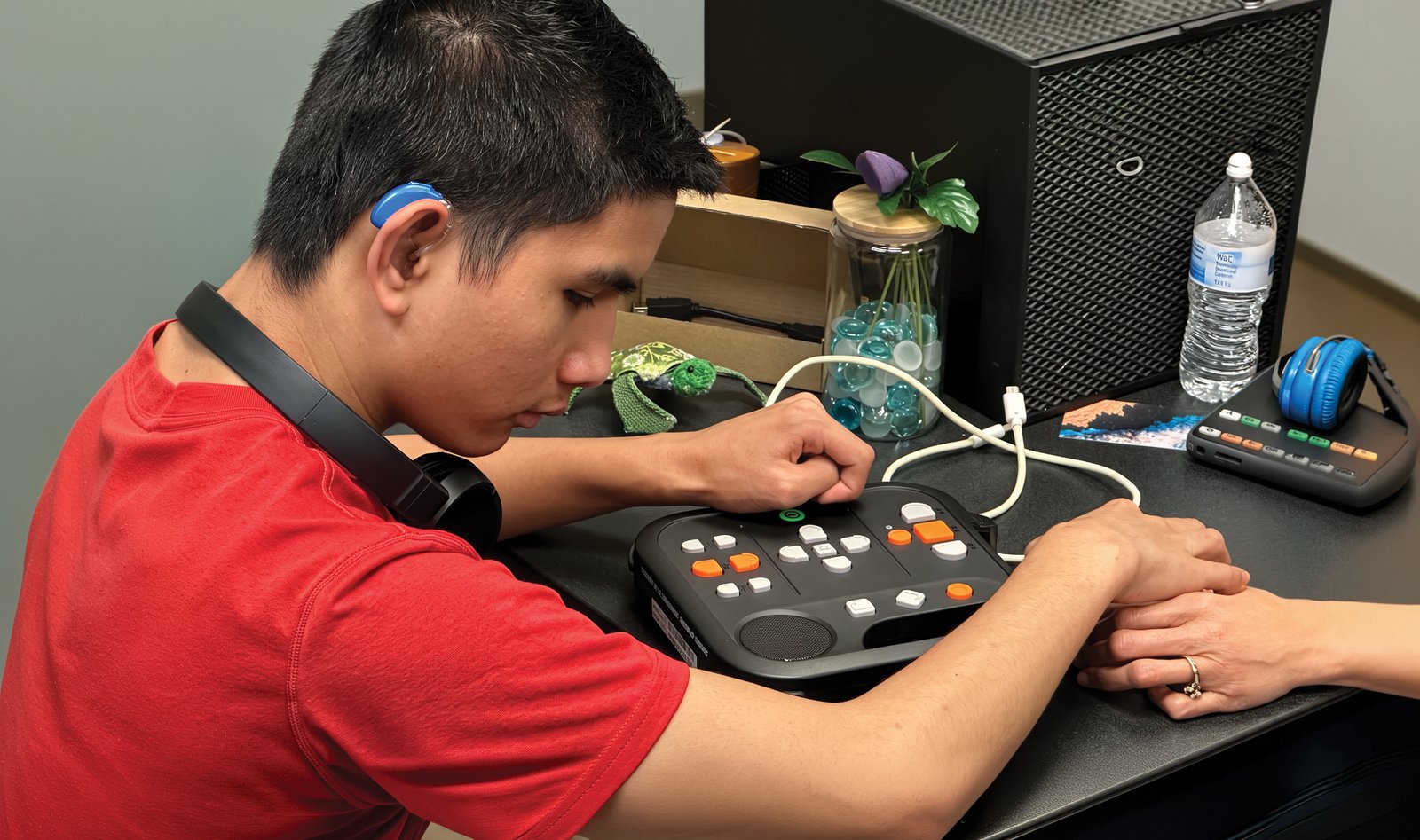 A young man wearing a hearing aid presses buttons on a tactile communication device at a desk while an instructor's hand rests beside his.