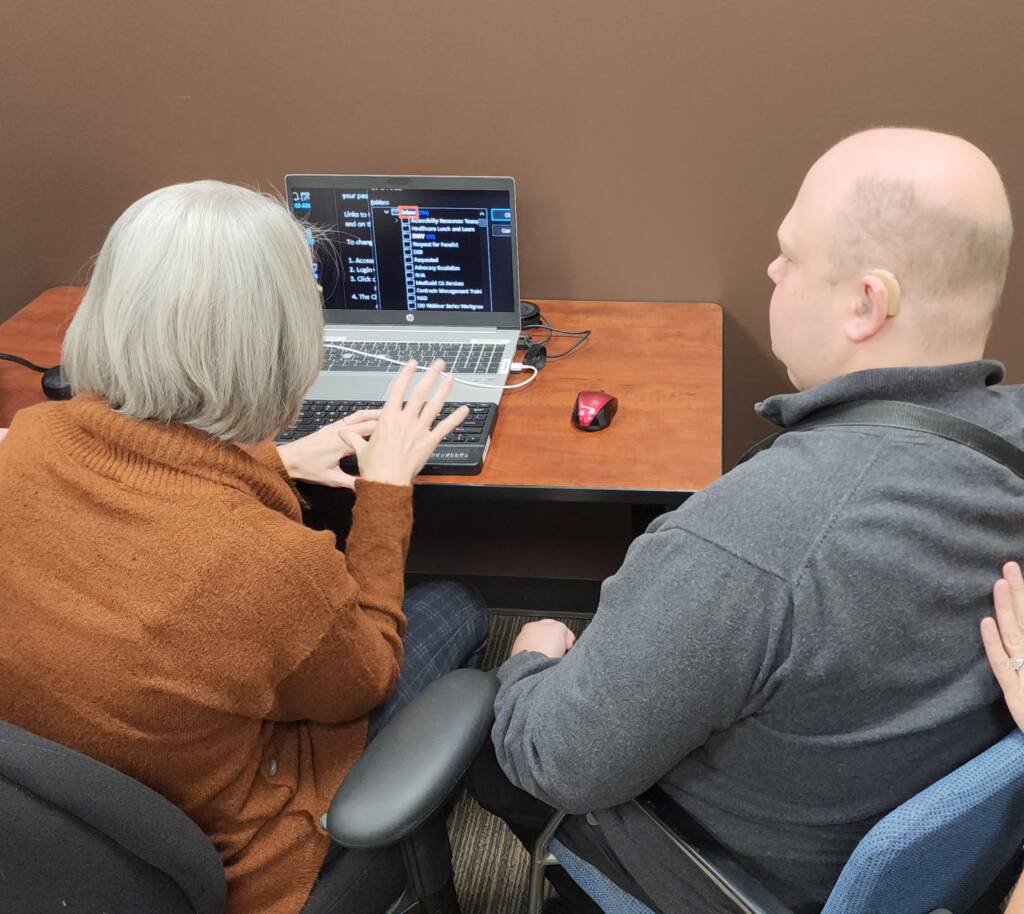 A woman with short grey hair and a brown top, sits at a desk while navigating a laptop with her braille display. Her instructor, a bald man in a grey shirt, sits to her right.