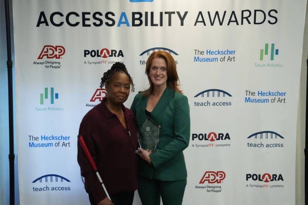 Two women stand together in front of the AccessAbility Awards backdrop, one holding an award and one holding a white cane.