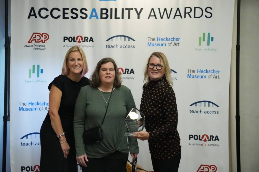 Three women stand together in front of the AccessAbility Awards backdrop, one holding an award.