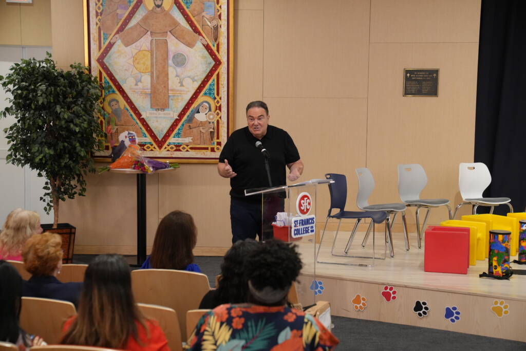 A man in a black polo shirt speaks from a podium at the front of the St. Francis College auditorium as seated guests listen.