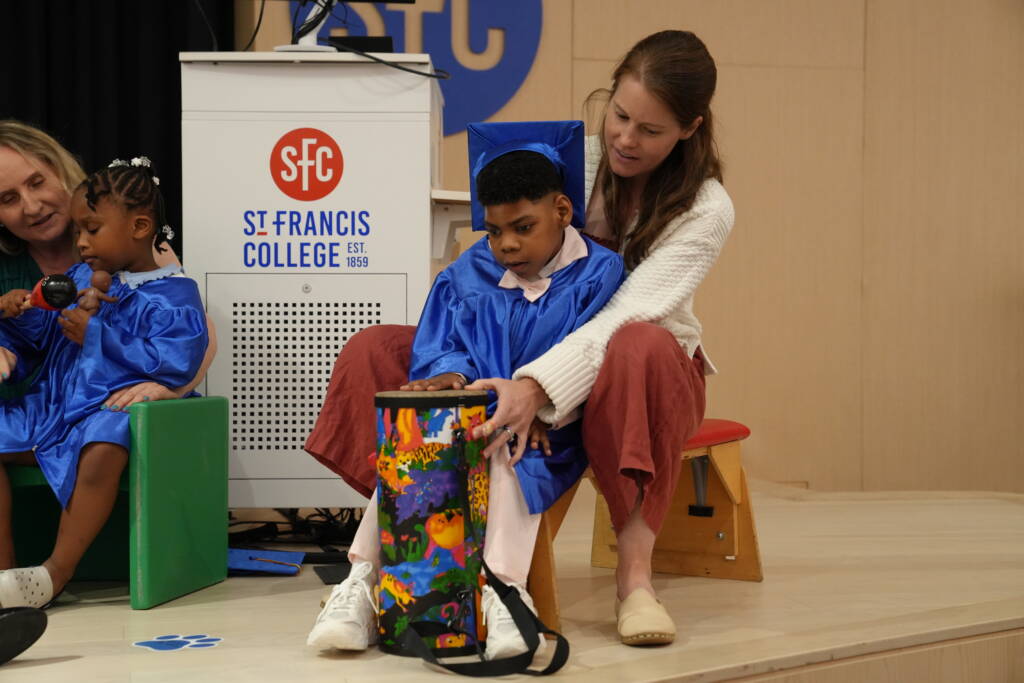 A young graduate in a blue cap and gown plays a colorful hand drum with help from a teacher seated behind him, while a classmate beside them shakes a maraca.