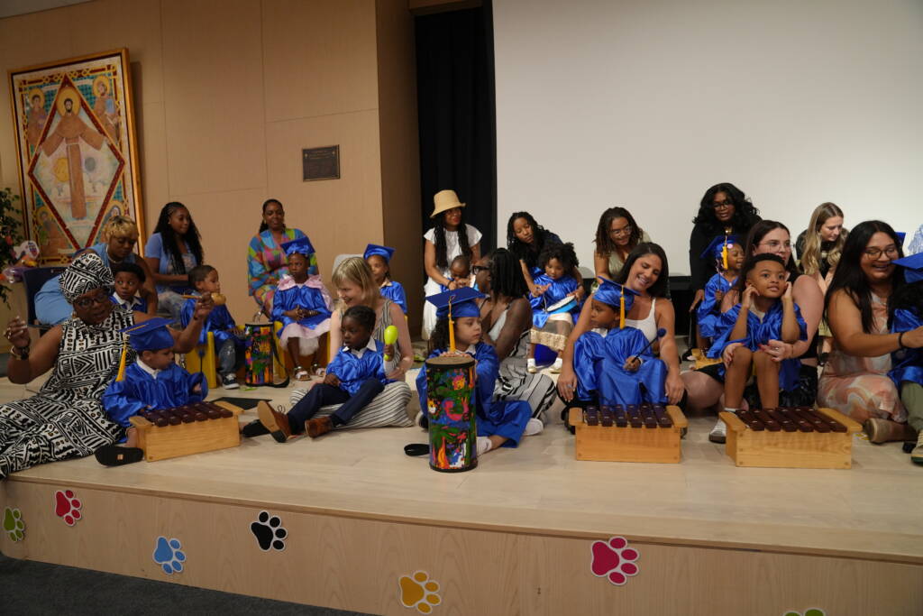 Graduates in blue caps and gowns sit along the edge of the stage with teachers and family members, playing xylophones, drums, and maracas together.