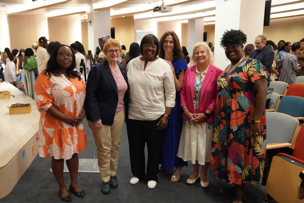 Six women stand together smiling for a group photo in the auditorium, with families and graduates gathered behind them.