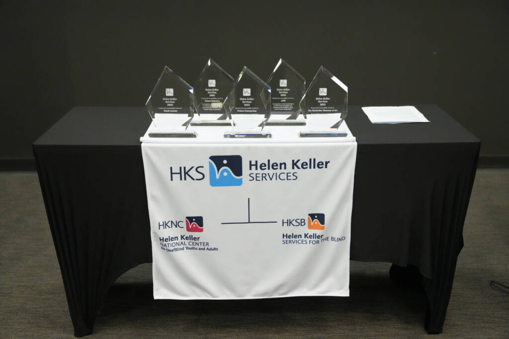 Five AccessAbility Awards lined up on a table draped with a Helen Keller Services banner.
