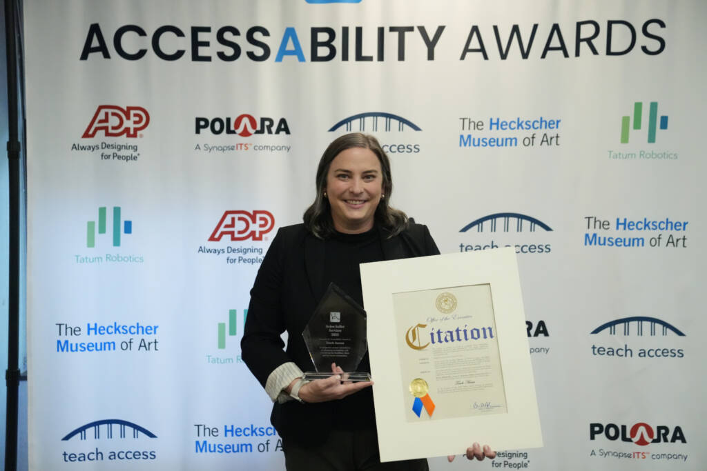 A woman stands in front of the AccessAbility Awards backdrop holding an award and a framed Nassau County citation.