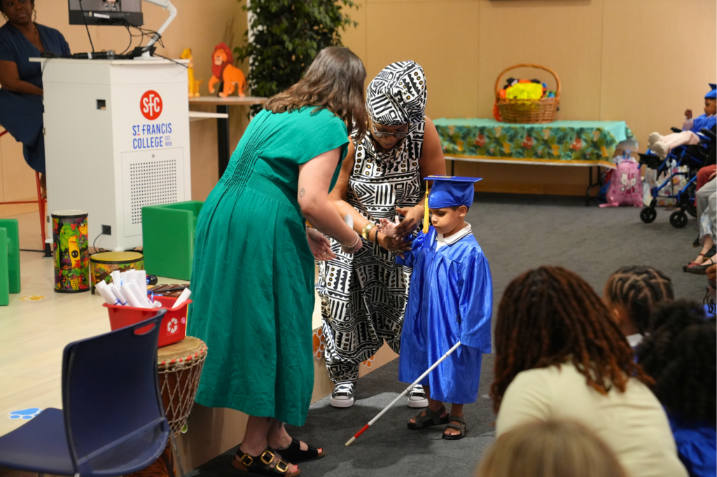 A young graduate in a blue cap and gown walks forward using a white cane, guided by two teachers, as guests watch from their seats.