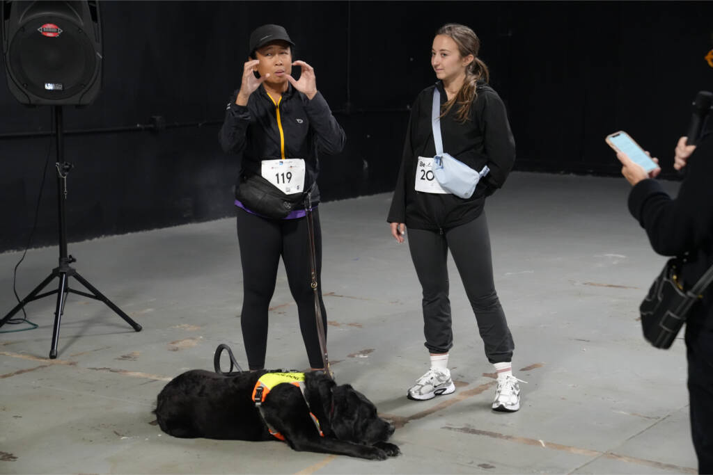 Maricar Marquez, race bib 119, signs beside her support service provider while her guide dog Cliff rests at their feet.