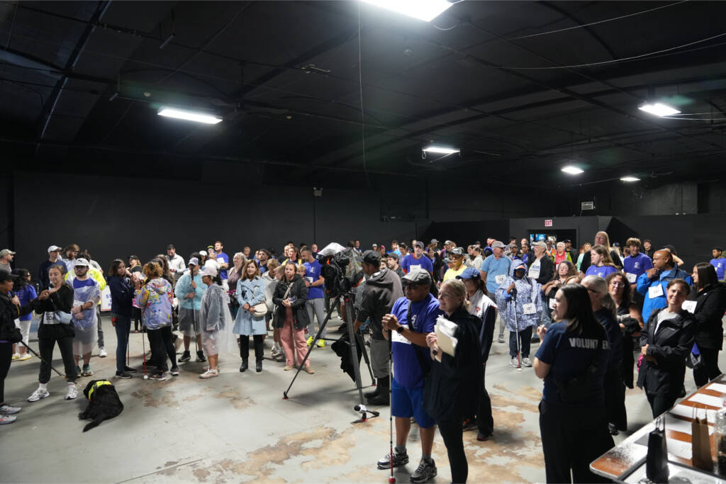 Participants fill the indoor gathering space before the race, with a News 12 camera set up in the center and a man in a purple event shirt holding a white cane in front.