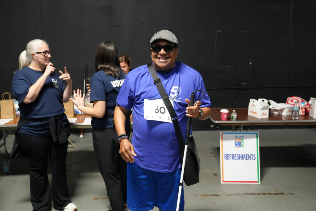 A smiling participant in a purple Helen's 5K shirt and bib 180 holds his white cane near the refreshments table while volunteers sign behind him.