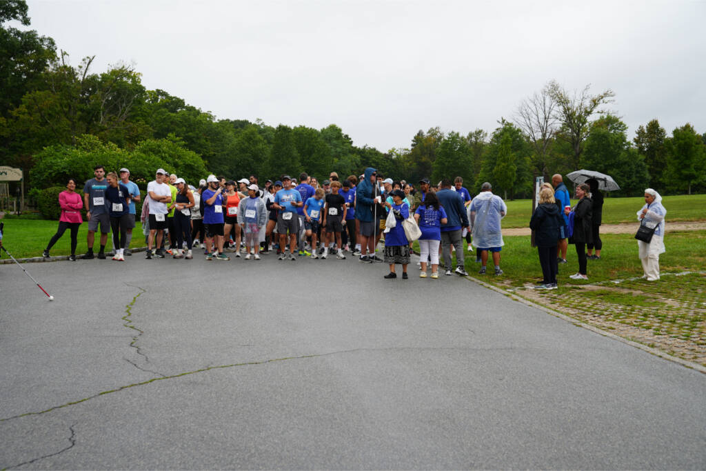 Walkers and runners gather near the start on a gray morning, some in rain ponchos, with a white cane in the foreground.