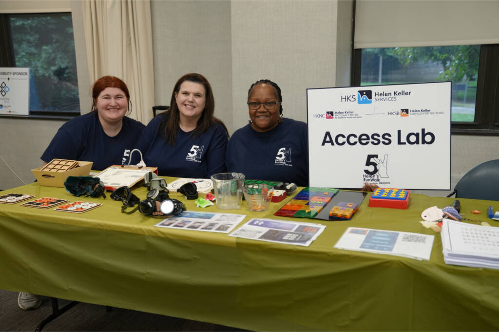 Three team members in Helen's RunWalk shirts sit behind the Access Lab table of tactile games, simulation goggles, and braille materials.